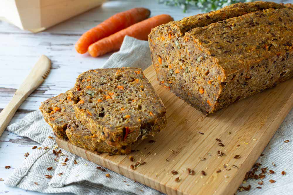 Dinkel-Karottenbrot auf einem Holzbrett, ein paar Scheiben sind schon abgeschnitten und liegen daneben. Im Hintergrund sind Möhren zu sehen.