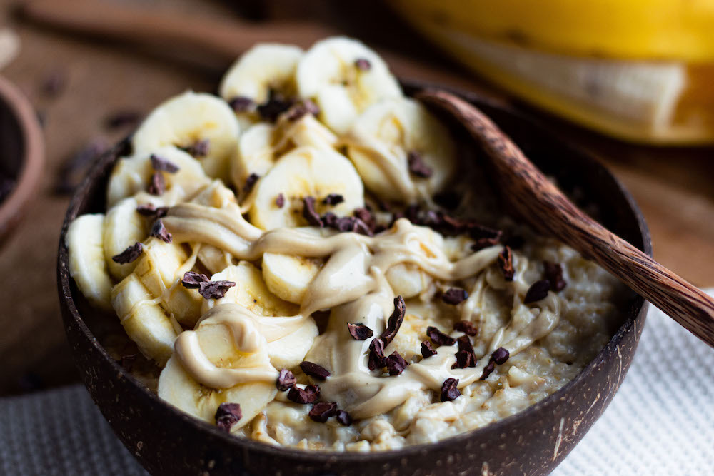Porridge in einer braunen Schüssel mit Bananenscheiben, Cashewmus und Kakaonibs dekoriert.