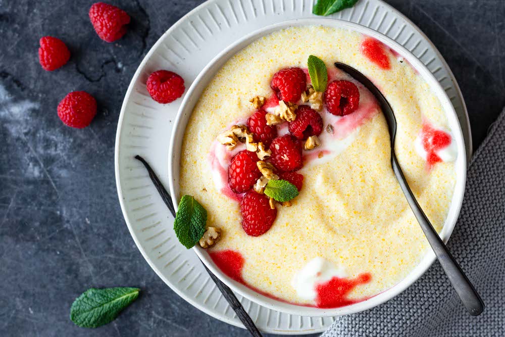 Polenta-Grießbrei in einer Schüssel serviert und dekoriert mit Himbeeren, Nüssen und Minze. Daneben liegt eine Vanilleschote.