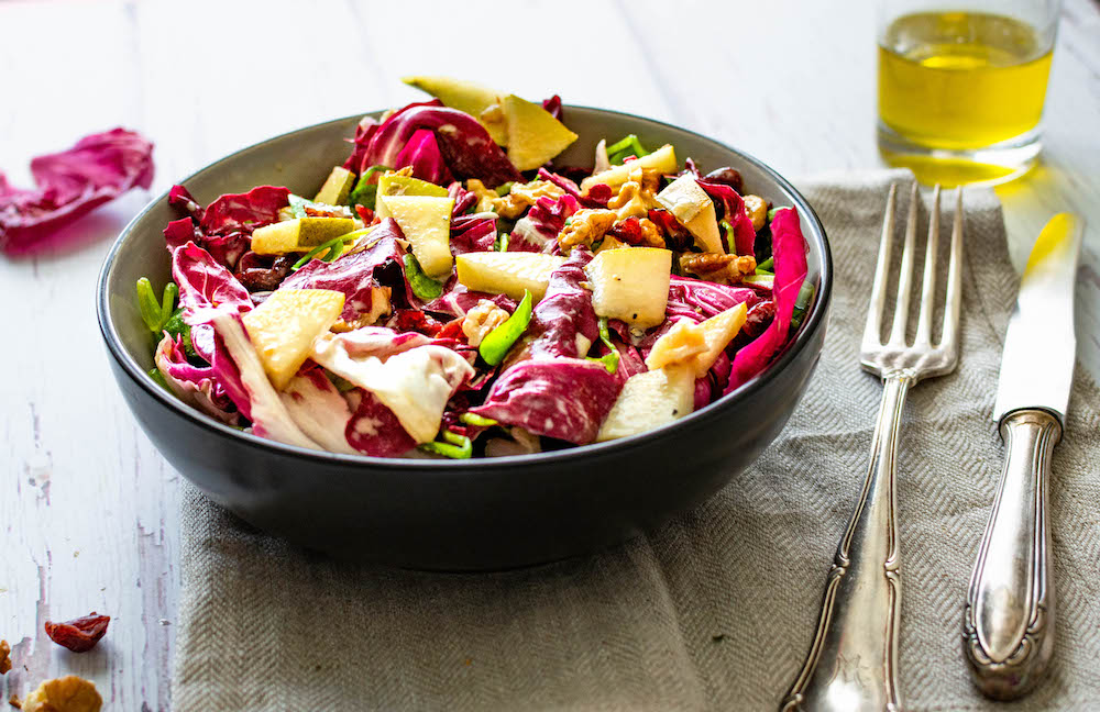 Postelein-Salat mit Radicchio, Birne, Walnüssen und Cranberries in einer Schüssel serviert, daneben liegt eine Gabel, im Hintergrund steht etwas Öl.