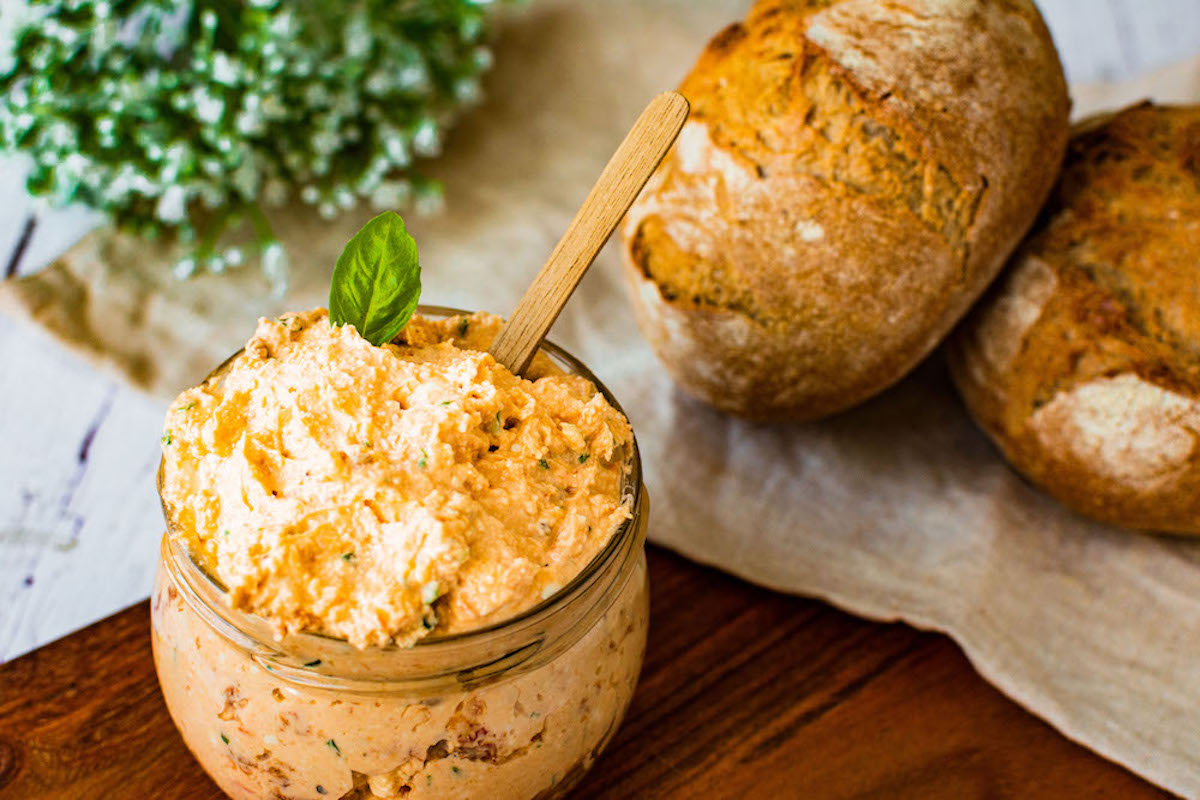 Feta-Quark-Dip in einem kleinen Glas, dekoriert mit einem Basilikumblatt. Ein kleiner Holzlöffel steckt im Dip und im Hintergrund liegen Brötchen.