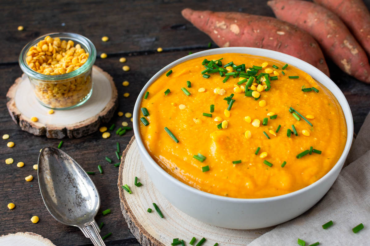 Gelb-orange Süßkartoffel-Linsen-Suppe in einer weißen Schüssel, mit Frühlingszwiebeln bestreut, daneben Süßkartoffeln und gelbe Linsen in einem Glas zur Deko.