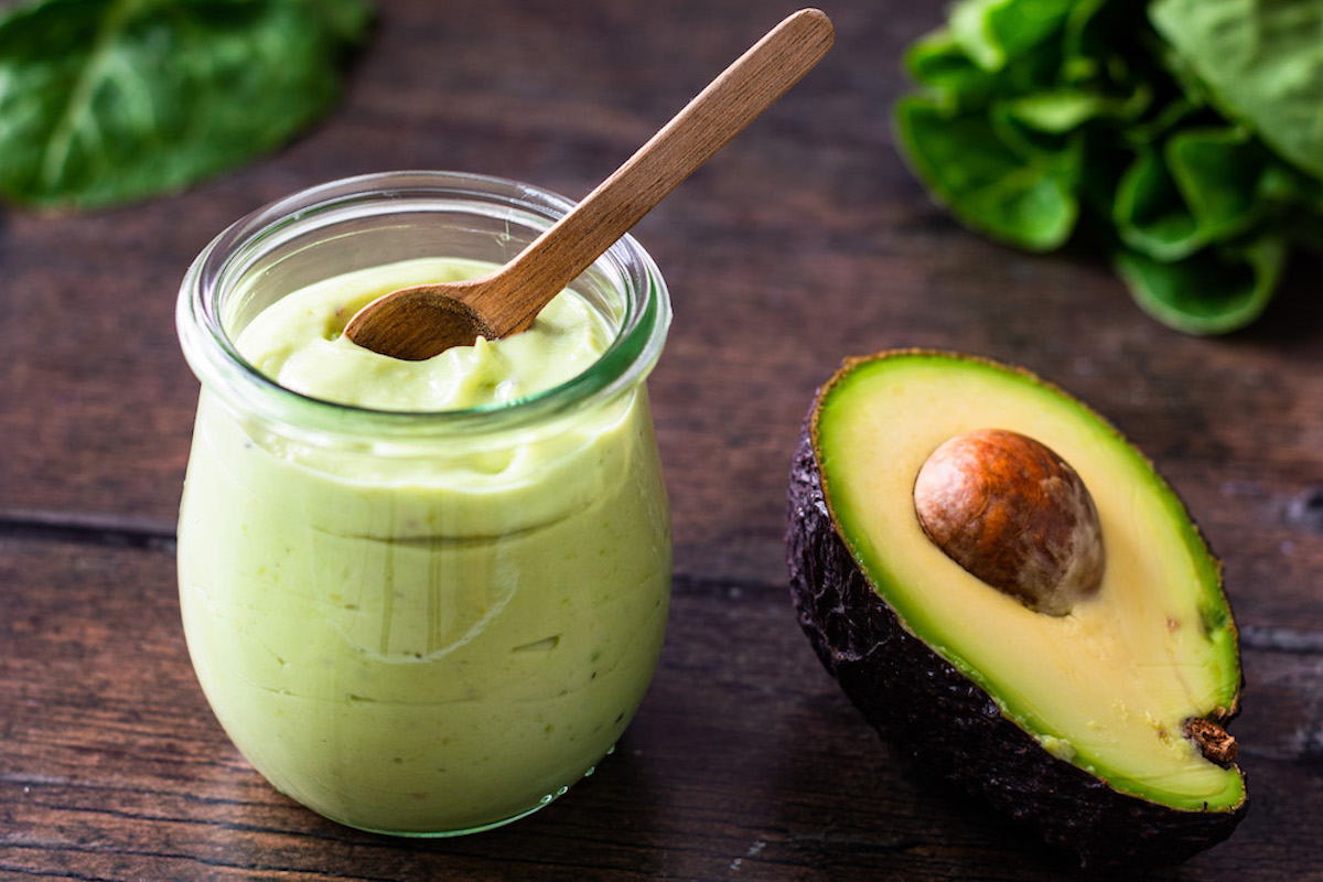Grünliches Dressing aus Avocado und Joghurt, welches in einem Weckglas serviert ist, in dem ein kleiner Holzlöffel steckt. Daneben liegt eine Avocado-Hälfte.