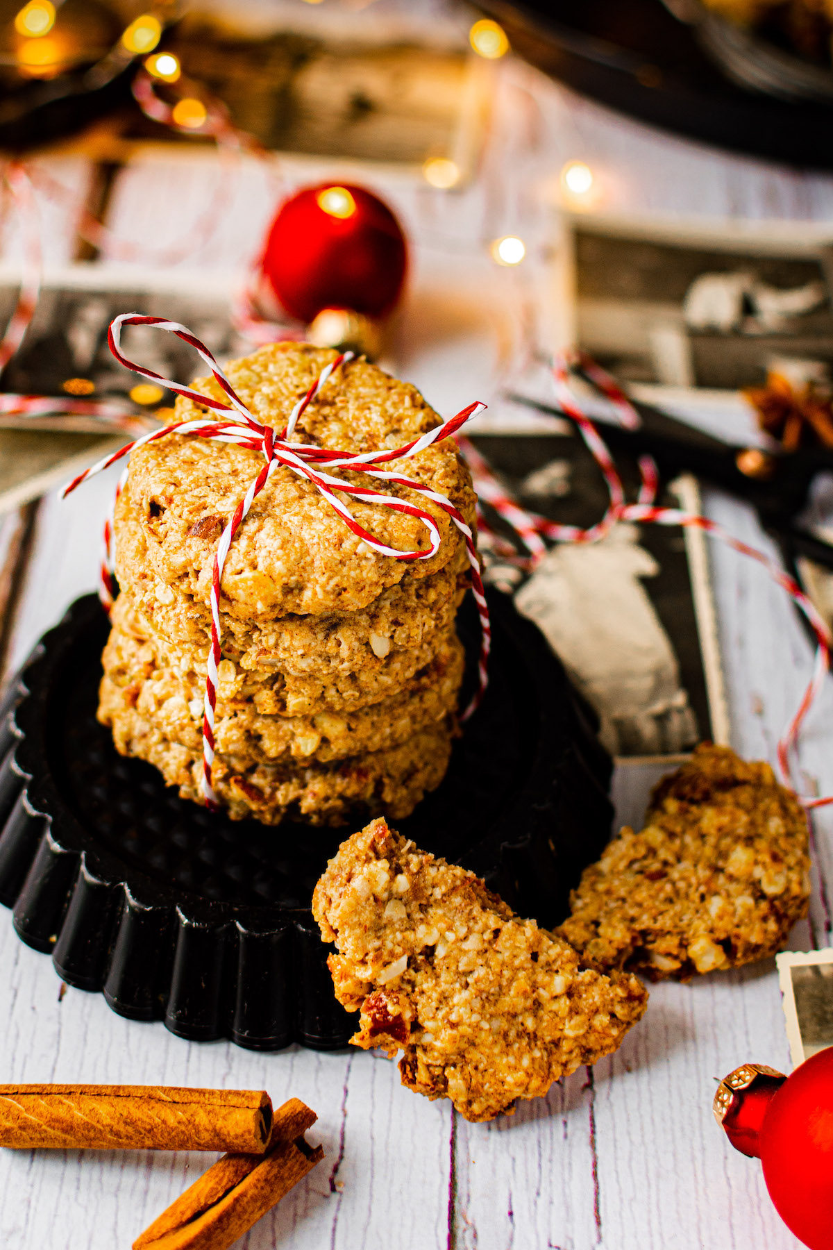 Weihnachtliche Cookies gestapelt und mit Geschenkband dekoriert. Drumherum liegen Zimtstangen, rote Christbaumkugeln, alte Bilder und eine Lichterkette.