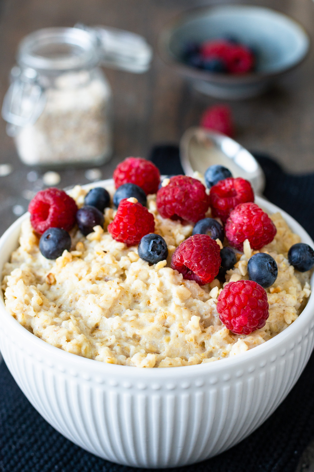 Selbstgemachter Porridge in einer weißen Schüssel mit Beeren dekoriert, im Hintergrund sind weitere Beeren und ein Glas mit Haferflocken zu sehen.