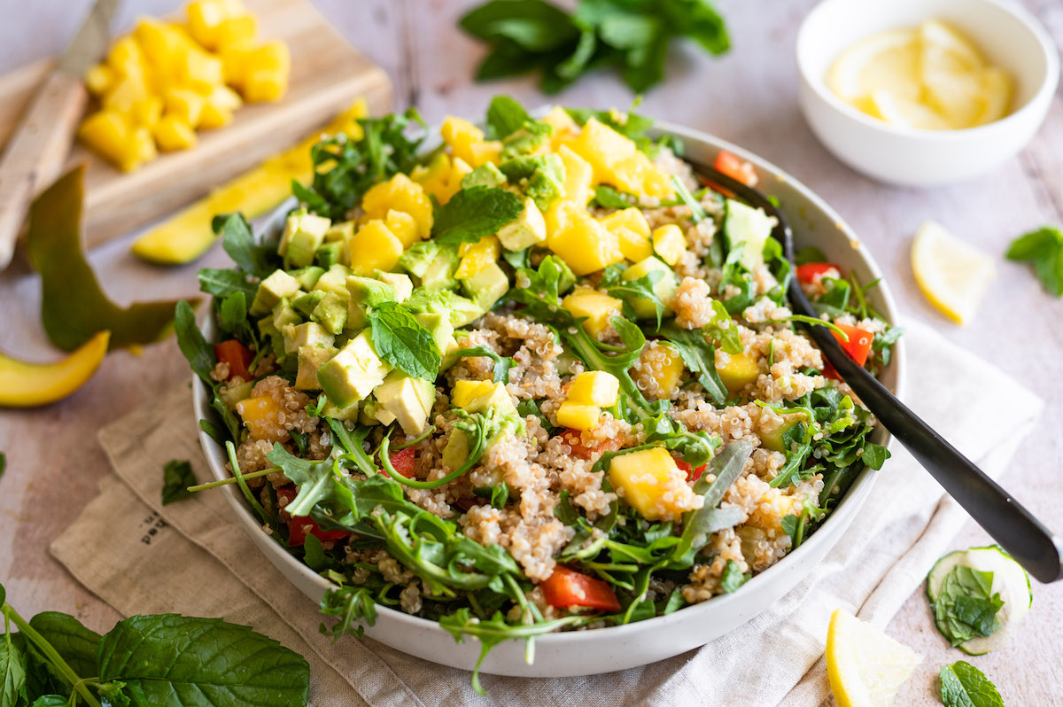 Quinoa-Salat mit Avocado und Mango, sowie Gurke, Paprika, Tomaten, Rucola und Minze. In einer weißen Schüssel serviert, in welcher eine schwarze Gabel steckt.