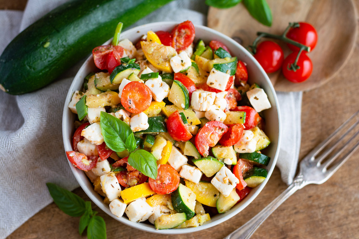 Salat mit Zucchini, Tomaten, Feta und Basilikum in einer Schüssel, daneben sind Zucchini, Tomaten und eine Gabel dekoriert.