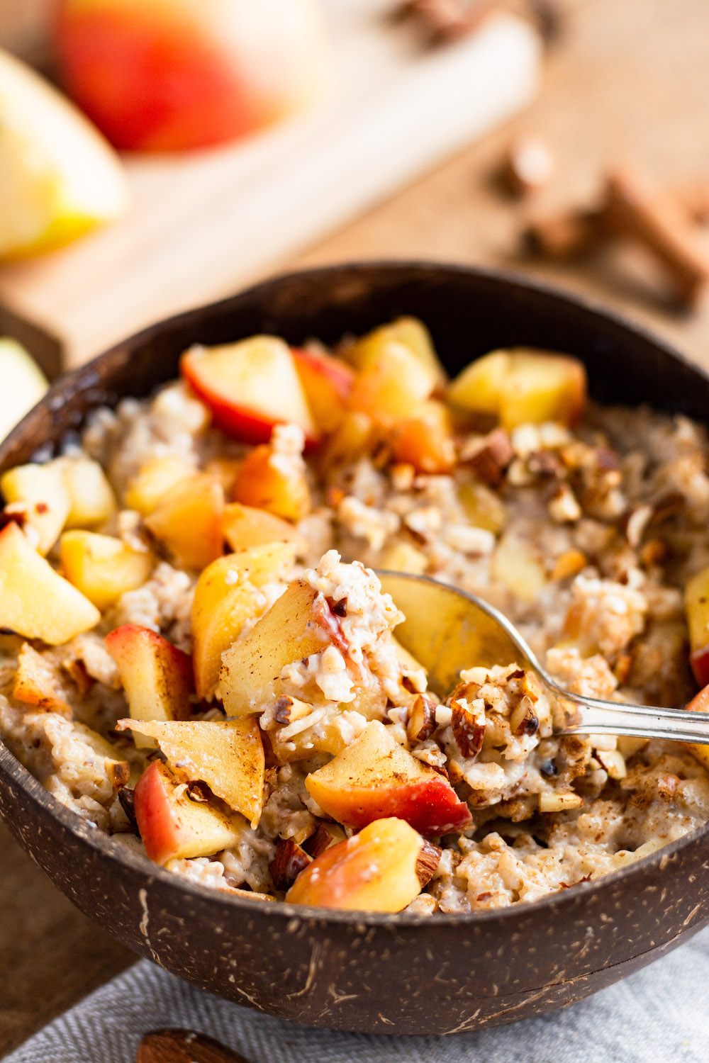 Apfel-Zimt-Porridge in einer Kokosschalenschüssel serviert und mit Zimtäpfeln und Mandelstücken garniert. Ein Löffel steckt im Haferbrei und dieser ist schon etwas vermischt.