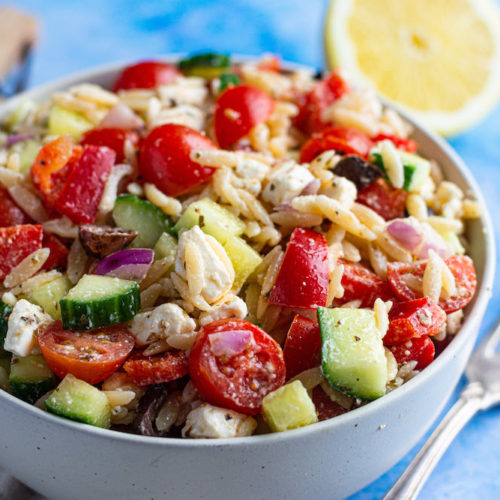 Griechischer Nudelsalat mit Kritharaki, Tomaten, Gurke und Feta in einer Schüssel serviert, daneben liegen eine Gabel und eine halbe Zitrone.