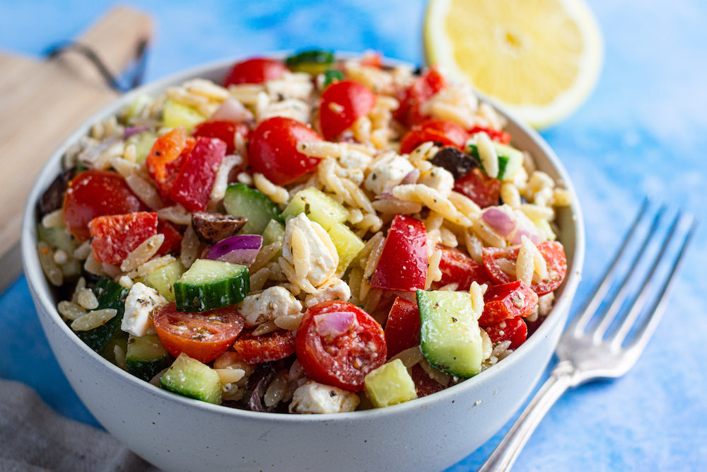 Griechischer Nudelsalat mit Kritharaki, Tomaten, Gurke und Feta in einer Schüssel serviert, daneben liegen eine Gabel und eine halbe Zitrone.