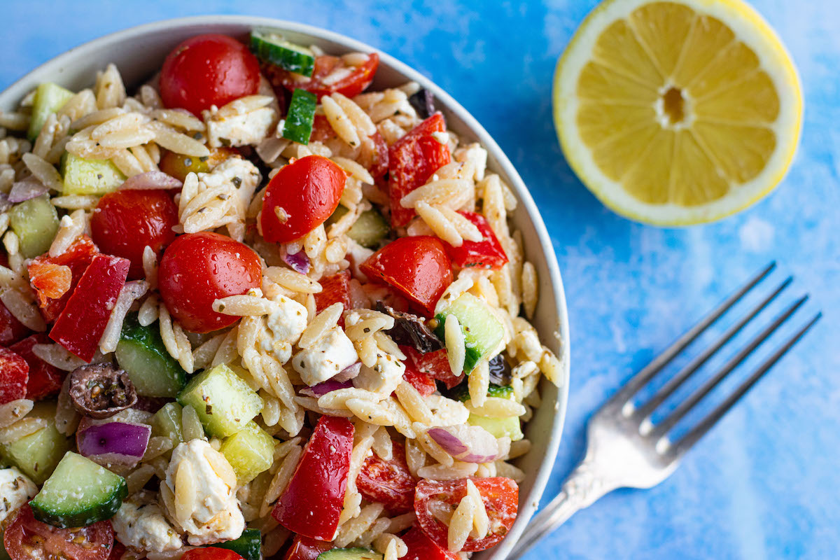 Griechischer Nudelsalat mit Kritharaki, Paprika, Tomaten, Gurke, Feta und Zwiebeln in einer Schüssel serviert, daneben liegen eine Gabel und eine halbe Zitrone.