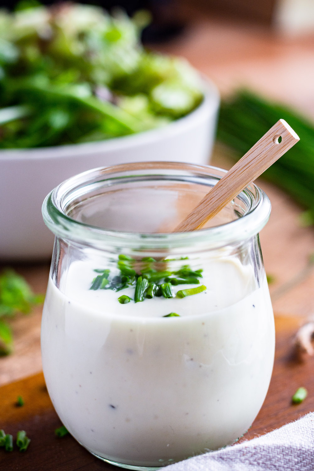 Joghurt-Senf-Dressing in einem Glas, in welchem ein kleiner Holzlöffel steckt. Mit Schnittlauch bestreut. Im Hintergrund steht eine Schüssel mit Salat.