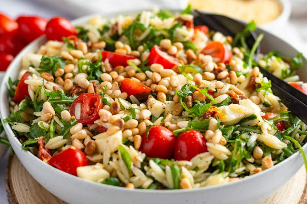 Orzo-Salat mit Tomaten, getrockneten Tomaten, Mozzarella und Rucola in einer weißen Schüssel mit Besteckt serviert.