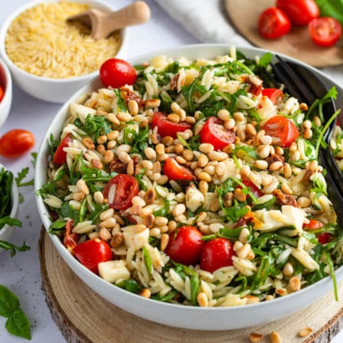 Mediterraner Orzo-Salat in weißer Schüssel serviert, in welcher schwarzes Besteck steckt.