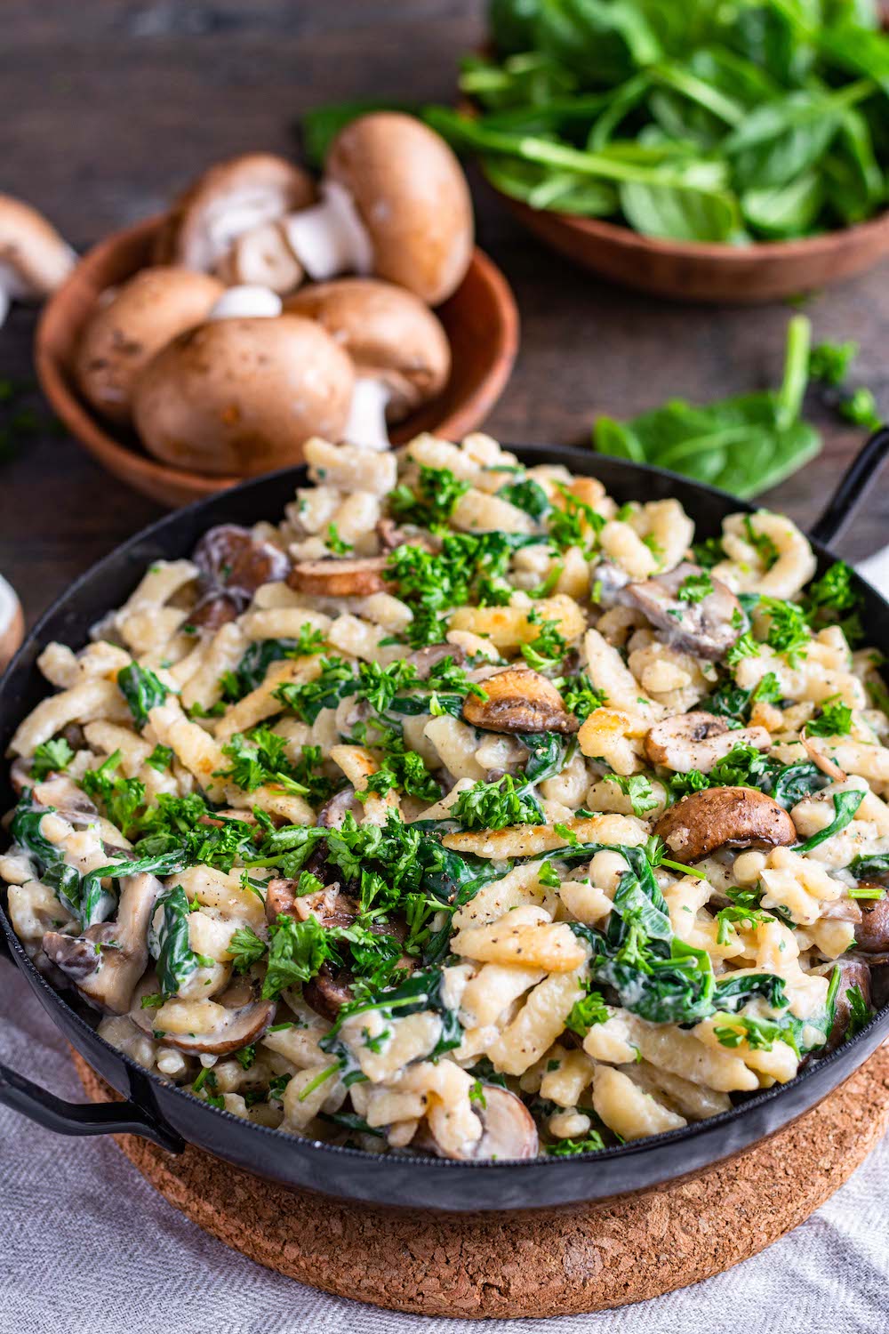 Schnelle Spätzlepfanne mit Spinat und Champignons in einer schwarzen Pfanne serviert, die auf einem Kork-Untersetzer steht.