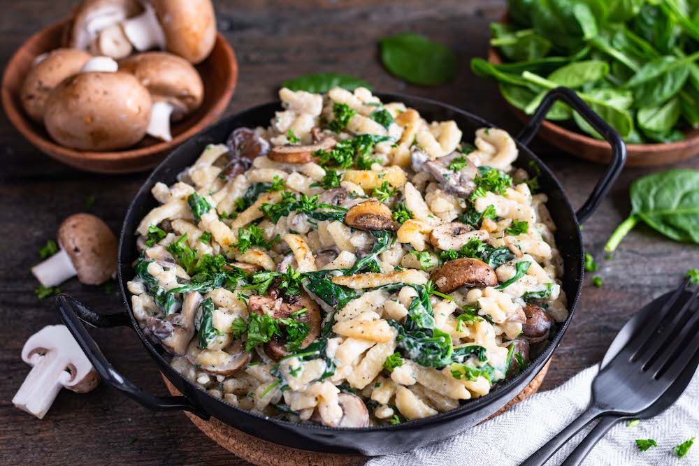 Spätzlepfanne mit Champignons und Spinat in einer schwarzen Pfanne mit Griffen serviert, im Hintergrund sind Champignons und Blattspinat dekoriert.