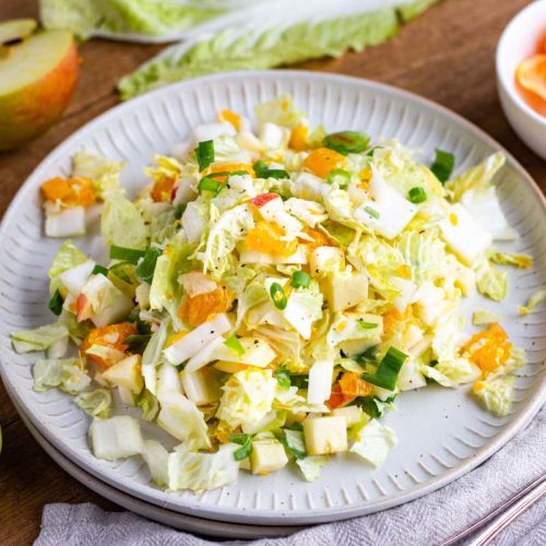 Chinakohl-Salat mit Apfel, Mandarinen und Frühlingszwiebeln auf einem matt weißen Teller serviert.