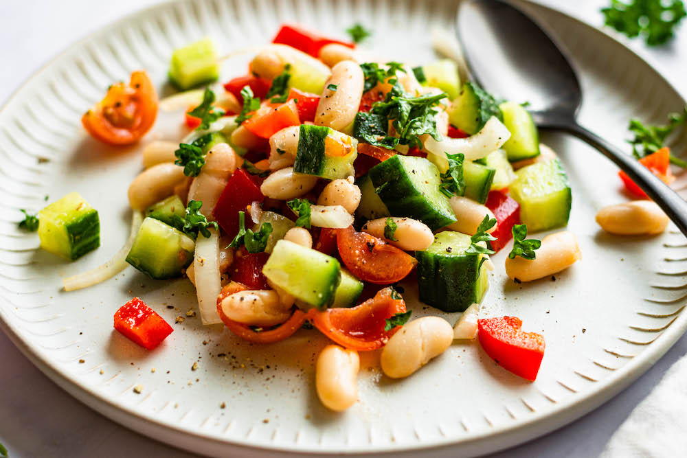 Weiße-Bohnen-Salat mit Gurke, Tomate, Paprika und Zwiebeln auf einem matt weißen Teller serviert.