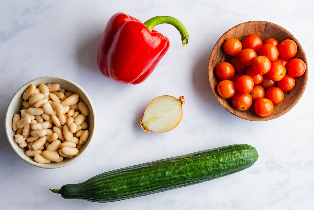 Die Zutaten für den weiße Bohnen Salat auf weißem Untergrund: Eine rote Paprika, weiße Bohnen aus der Dose, eine halbe Zwiebel, Cherrytomaten in einer Schüssel und eine Salatgurke.