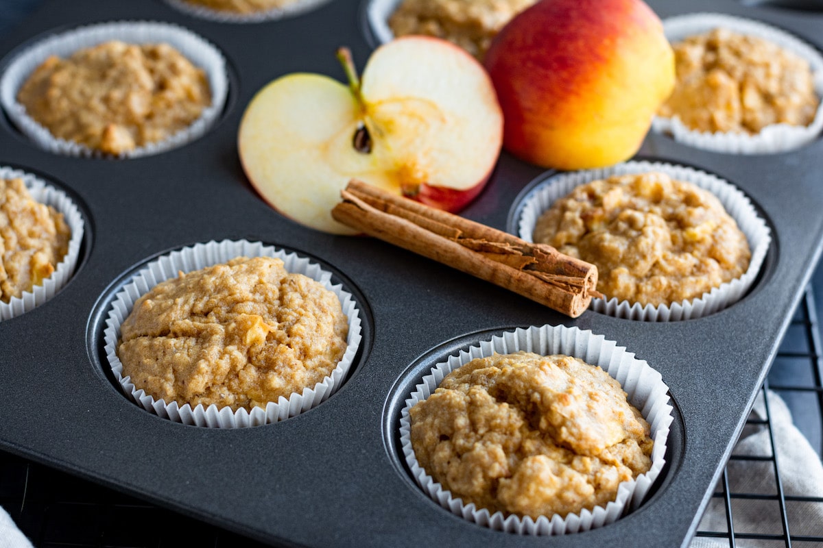 Gebackene Apfelmuffins in einem Muffinsblech, auf welchem außerdem ein angeschnittener Apfel und eine Zimtstange liegen.