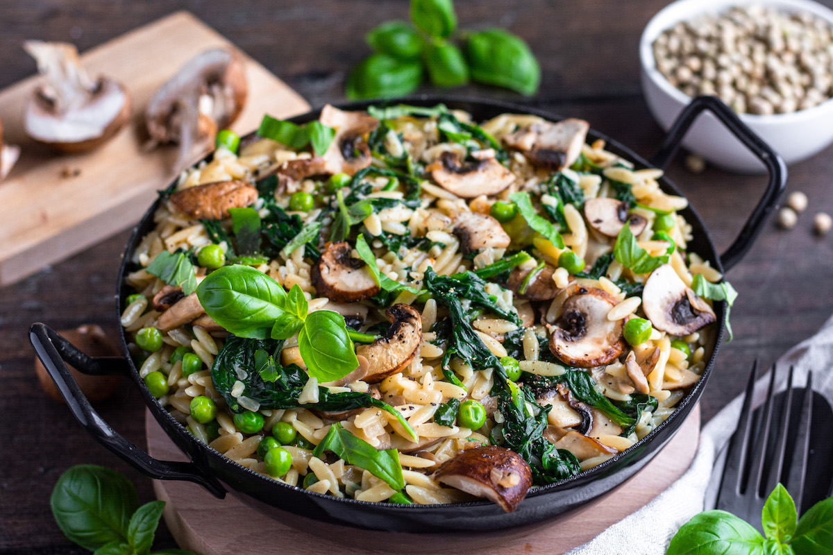 Orzo-Risotto mit Spinat, Champignons und Erbsen in einer schwarzen Pfanne serviert und mit Basilikum dekoriert.