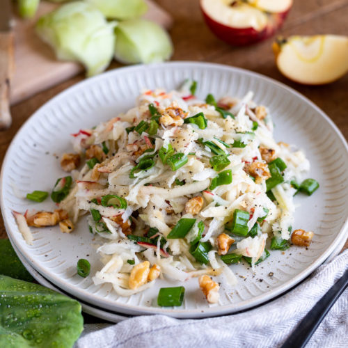 Geraspelter Kohlrabisalat mit Apfel, Frühlingszwiebeln und Walnüssen auf einem flachen Teller angerichtet.