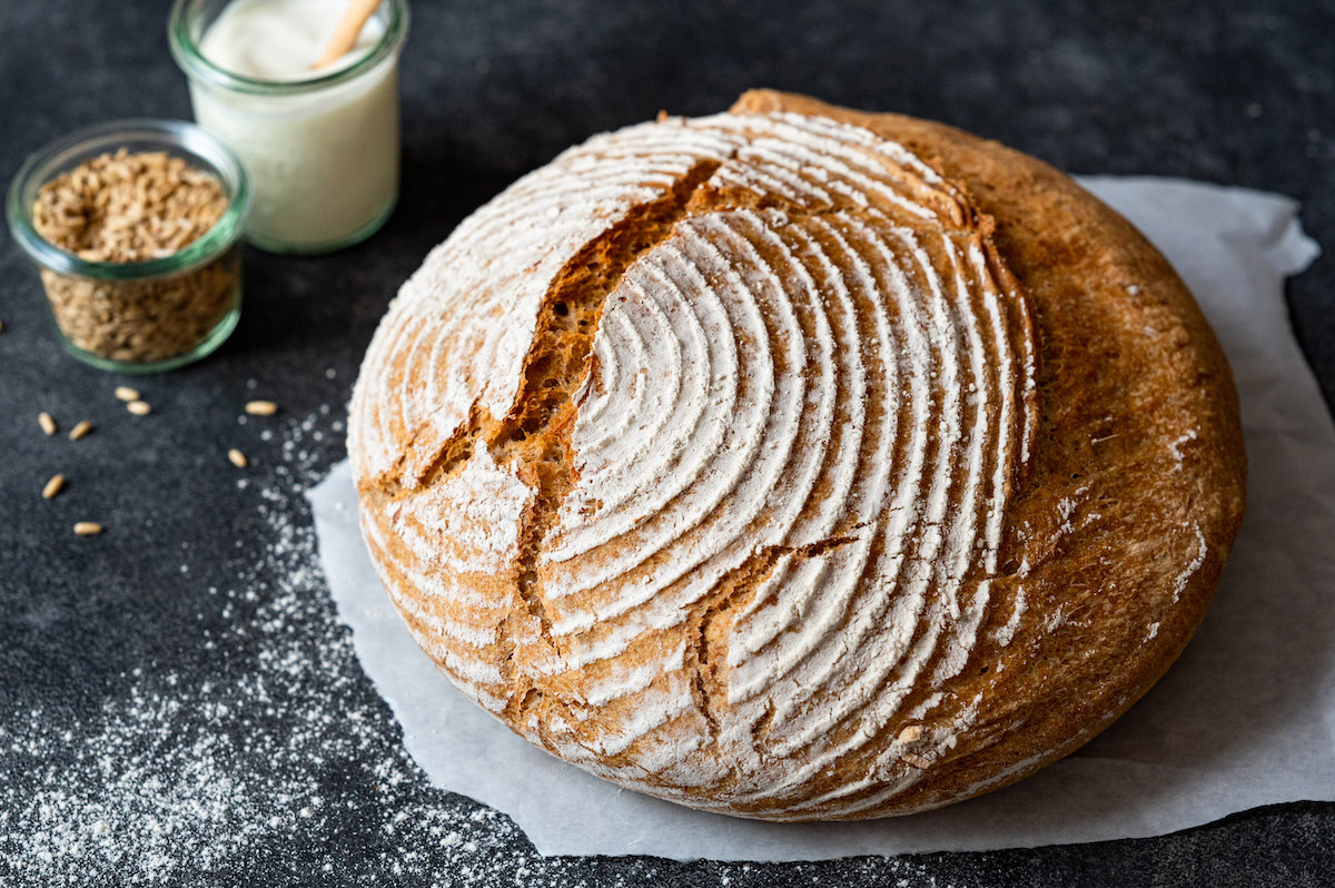 Dinkel-Joghurt-Brot mit aufgerissener Kruste auf einem weißen Backpapier auf schwarzem Untergrund.