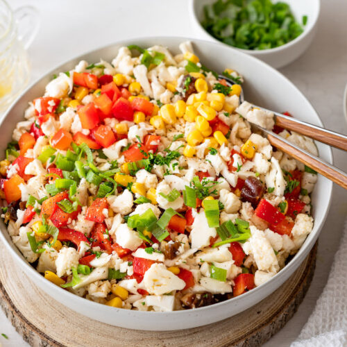 Roher Blumenkohlsalat mit Paprika, Mais und Frühlingszwiebeln in einer weißen Schüssel serviert.