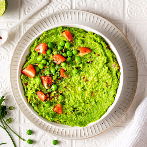 Erbsen-Guacamole mit Tomaten, Frühlingszwiebeln und Petersilie dekoriert in einer weißen Schüssel von oben fotografiert.