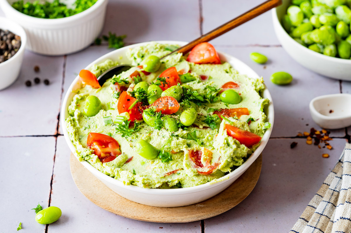 Edamole (Edamame Dip) in einer kleinen Schüssel serviert und mit Cherrytomaten, Petersilie und Frühlingszwiebeln, sowie ganzen Edamame Bohnen dekoriert.