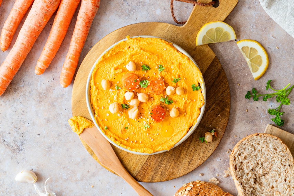 Karotten-Hummus in einer Schüssel angerichtet, mit Kichererbsen, Möhrenstücken und Sesam dekoriert.