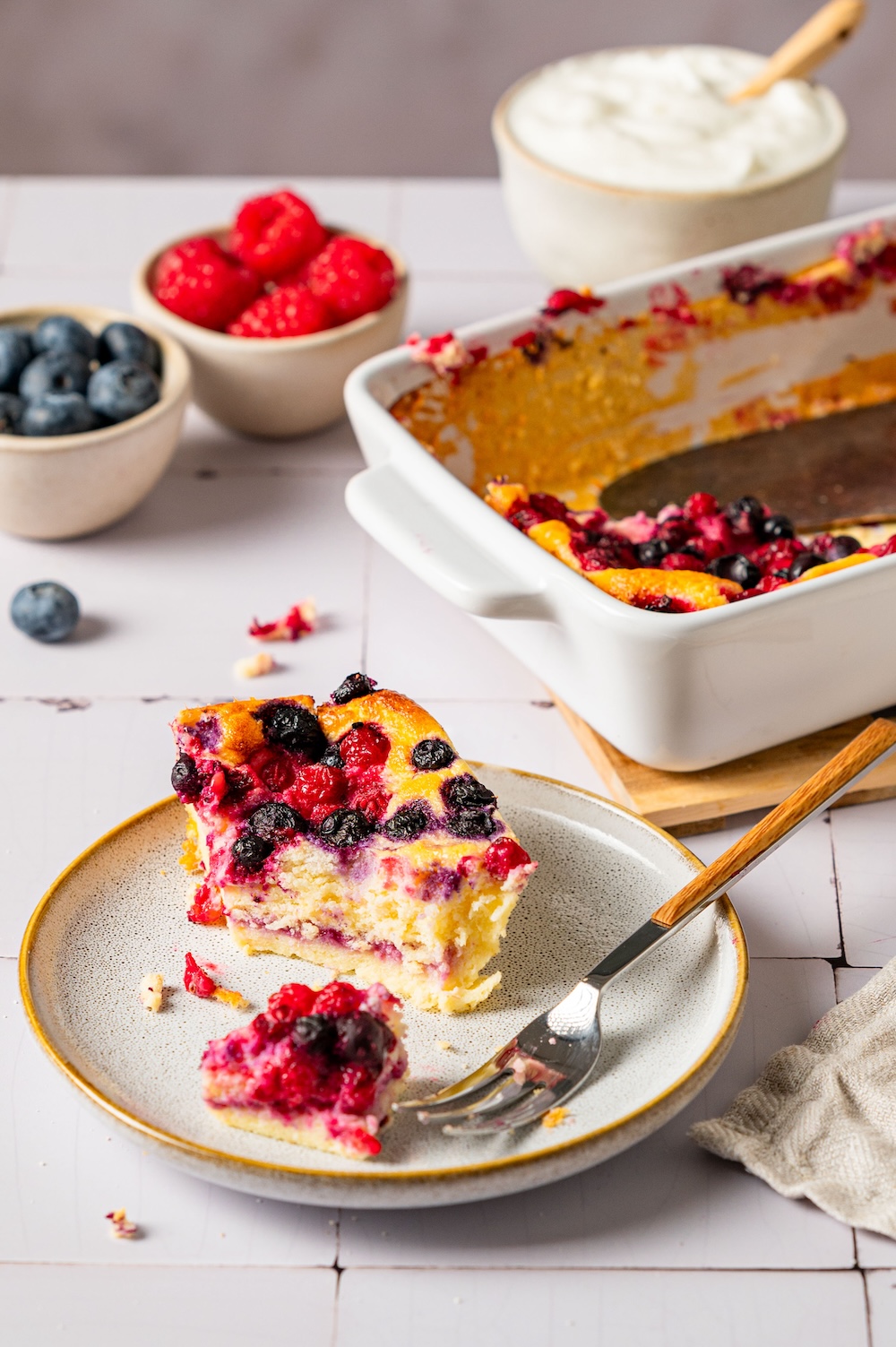 Ein Stück Skyr Kuchen auf einem Teller, im Hintergrund ist die Auflaufform zu sehen.
