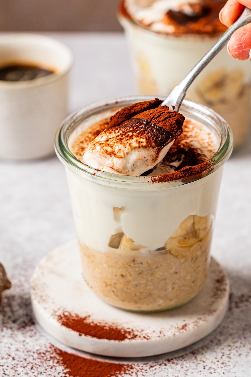 Nahaufnahme von Tiramisu Overnight Oats im Glas. Ein Stück der Skyr Schicht mit Kakaopulver wird gerade von einem Löffel herausgenommen.