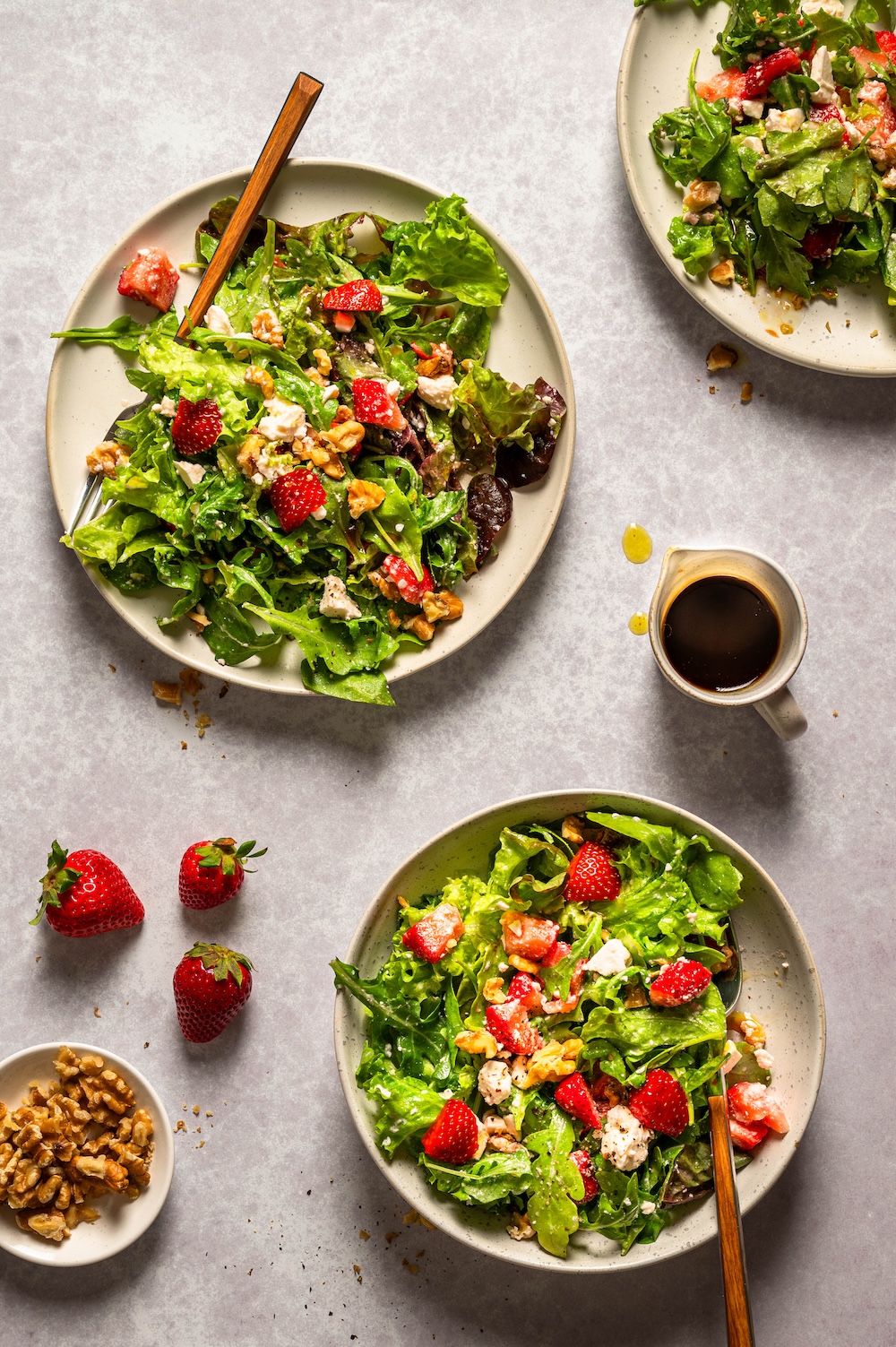 Sommerlicher Salat mit Erdbeeren, Rucola, Feta und Walnüssen auf 3 Tellern bzw. Schüsseln angerichtet.