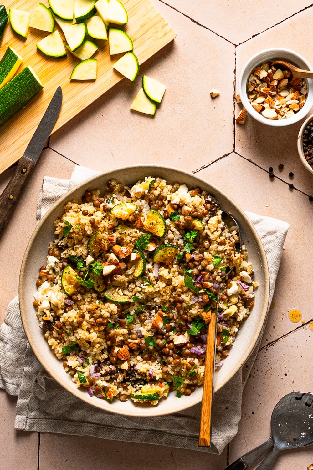 Bulgursalat mit Linsen, Zucchini und Feta in einer Schüssel. Daneben ein Schneidbrett mit geschnittener Zucchini.