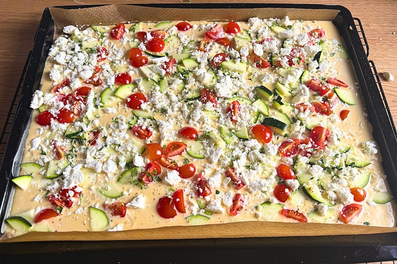Blech mit Teig befüllt und mit Zucchini, Tomaten und Feta als Ofenpfannkuchen belegt.