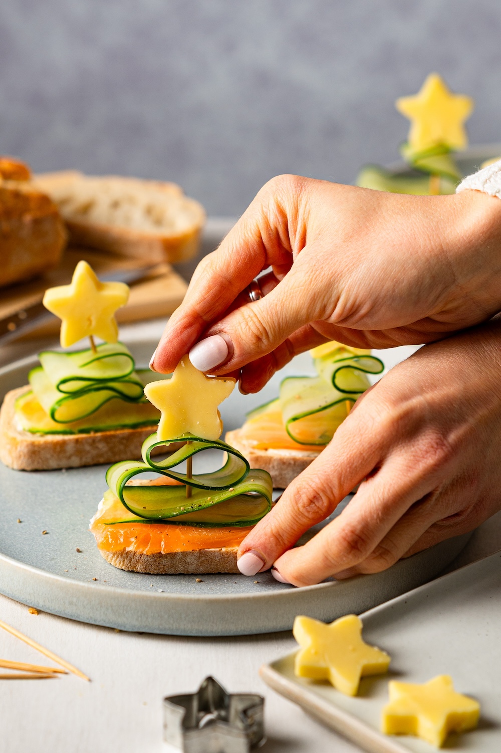 Brote mit Räucherlachs und Gurken-Tannenbaum-Spießen, auf welche gerade ein Stern aus Käse gesteckt wird.