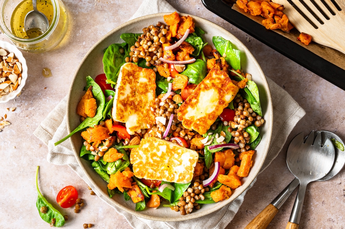Süßkartoffel-Linsen-Salat mit Spinat, Zwiebeln, Tomaten, Mandeln und gebratenem Halloumi in einer Schüssel serviert.